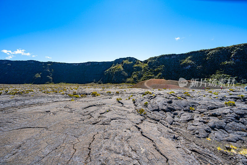 游客在凝固的熔岩场中间的自然火山景观中探索一个小火山口，背景是一个巨大的绿色覆盖的火山口边缘。Formica Leo, Piton de la Fournaise，留尼旺岛，法国，欧洲。